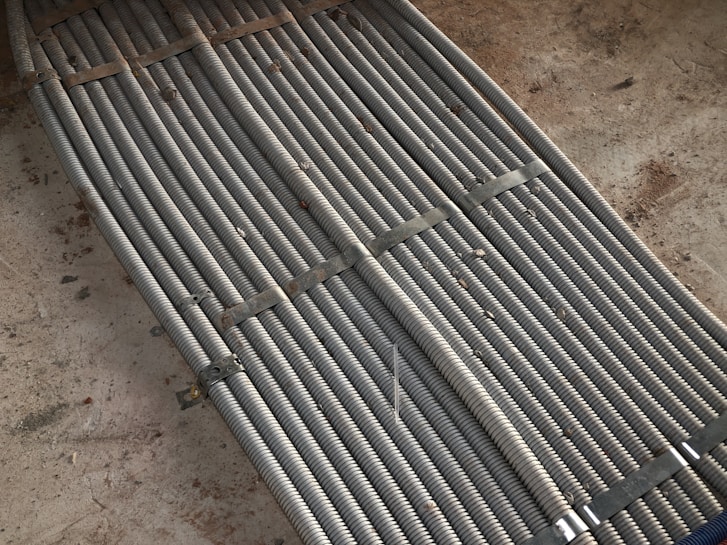 Metal coils are tightly bundled together, secured with brackets, forming a uniform and structured pattern. There is a background of rough concrete with scattered dirt and debris.