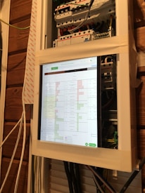 A close-up view of an electrical panel with a digital screen displaying a spreadsheet. The panel is secured with masking tape, and various colored wires are visible. The wooden wall in the background provides a rustic contrast to the modern equipment.