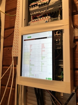 A close-up view of an electrical panel with a digital screen displaying a spreadsheet. The panel is secured with masking tape, and various colored wires are visible. The wooden wall in the background provides a rustic contrast to the modern equipment.
