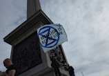 Several people stand at the base of a monument holding a white flag with a blue symbol and the text 'REBELLION IS ACT OF LOVE' against a cloudy sky.