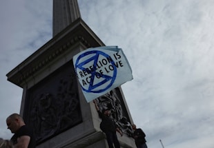 Several people stand at the base of a monument holding a white flag with a blue symbol and the text 'REBELLION IS ACT OF LOVE' against a cloudy sky.