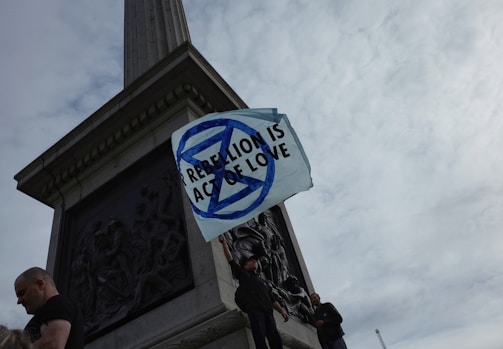 Several people stand at the base of a monument holding a white flag with a blue symbol and the text 'REBELLION IS ACT OF LOVE' against a cloudy sky.