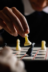 A person is holding a white chess piece, likely a rook, over a board in a strategic move. The focus is on the hand and the piece, while a black knight sits on the board. Other chess pieces are blurred in the background.