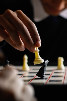 A person is holding a white chess piece, likely a rook, over a board in a strategic move. The focus is on the hand and the piece, while a black knight sits on the board. Other chess pieces are blurred in the background.