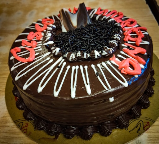 A round chocolate cake decorated with chocolate shavings and white icing lines. Red icing spells out a message on top. The cake is set on a simple background, likely a wooden table.