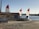 A sandy beach area featuring two sailboats with red, white, and blue sails near a small rental shack. The shack has a sign reading 'THE SANDBOX BIKE RENTAL.' A billboard advertises 'Hurricane Boat Rentals' beside it. The scene is set against a calm body of water, with trees and amusement park rides visible in the distance.