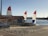 A sandy beach area featuring two sailboats with red, white, and blue sails near a small rental shack. The shack has a sign reading 'THE SANDBOX BIKE RENTAL.' A billboard advertises 'Hurricane Boat Rentals' beside it. The scene is set against a calm body of water, with trees and amusement park rides visible in the distance.