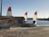 A sandy beach area featuring two sailboats with red, white, and blue sails near a small rental shack. The shack has a sign reading 'THE SANDBOX BIKE RENTAL.' A billboard advertises 'Hurricane Boat Rentals' beside it. The scene is set against a calm body of water, with trees and amusement park rides visible in the distance.