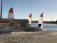 A sandy beach area featuring two sailboats with red, white, and blue sails near a small rental shack. The shack has a sign reading 'THE SANDBOX BIKE RENTAL.' A billboard advertises 'Hurricane Boat Rentals' beside it. The scene is set against a calm body of water, with trees and amusement park rides visible in the distance.