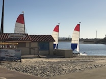 A sandy beach area featuring two sailboats with red, white, and blue sails near a small rental shack. The shack has a sign reading 'THE SANDBOX BIKE RENTAL.' A billboard advertises 'Hurricane Boat Rentals' beside it. The scene is set against a calm body of water, with trees and amusement park rides visible in the distance.
