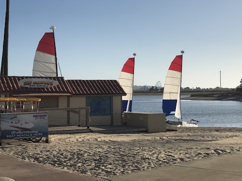 A sandy beach area featuring two sailboats with red, white, and blue sails near a small rental shack. The shack has a sign reading 'THE SANDBOX BIKE RENTAL.' A billboard advertises 'Hurricane Boat Rentals' beside it. The scene is set against a calm body of water, with trees and amusement park rides visible in the distance.