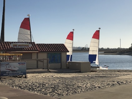 A sandy beach area featuring two sailboats with red, white, and blue sails near a small rental shack. The shack has a sign reading 'THE SANDBOX BIKE RENTAL.' A billboard advertises 'Hurricane Boat Rentals' beside it. The scene is set against a calm body of water, with trees and amusement park rides visible in the distance.