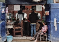 A small, rustic barbershop with two customers and a barber. The shop has two large mirrors and an array of shelves filled with various grooming products. There are towels hanging above the mirrors. One customer is sitting on a chair getting a haircut, while another person is sitting in the middle waiting. There's a child sitting on a chair by the entrance watching the scene.