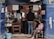 A small, rustic barbershop with two customers and a barber. The shop has two large mirrors and an array of shelves filled with various grooming products. There are towels hanging above the mirrors. One customer is sitting on a chair getting a haircut, while another person is sitting in the middle waiting. There's a child sitting on a chair by the entrance watching the scene.