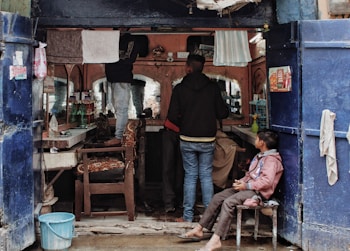 A small, rustic barbershop with two customers and a barber. The shop has two large mirrors and an array of shelves filled with various grooming products. There are towels hanging above the mirrors. One customer is sitting on a chair getting a haircut, while another person is sitting in the middle waiting. There's a child sitting on a chair by the entrance watching the scene.