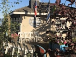 A small wooden building with a shingled roof, displaying a sign that reads 'Beach House'. Multiple flags are flying from the roof. White plastic chairs are arranged outside next to colorful flower planters. The area is surrounded by greenery and shadows cast by nearby plants.