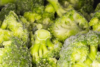 Close-up view of several broccoli florets covered with a layer of frost, indicating they are frozen. The surface texture of the broccoli is detailed, showing its bumpy and leafy parts.