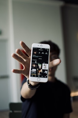 A person holds a smartphone displaying a social media profile with photos. The focus is on the phone with the individual's profile interface visible, set against a blurred background.