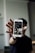 A person holds a smartphone displaying a social media profile with photos. The focus is on the phone with the individual's profile interface visible, set against a blurred background.