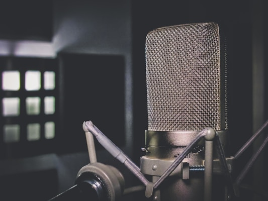 A close-up of a vintage-style microphone against a blurred background with geometric shapes. The microphone's metal grille and stand are prominently featured with a muted color palette.