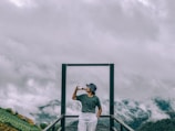 A person stands on a platform with a metal frame, wearing a hat and holding a cup. They are positioned against a backdrop of mountains partially covered with mist and clouds, creating a serene and tranquil atmosphere.