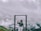 A person stands on a platform with a metal frame, wearing a hat and holding a cup. They are positioned against a backdrop of mountains partially covered with mist and clouds, creating a serene and tranquil atmosphere.