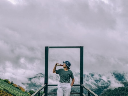 A person stands on a platform with a metal frame, wearing a hat and holding a cup. They are positioned against a backdrop of mountains partially covered with mist and clouds, creating a serene and tranquil atmosphere.