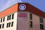 A building facade with a modern architectural design featuring a contrasting color scheme. The upper part of the structure is accented with a deep red section, prominently displaying a round emblem. Below, the words 'Universidad Metropolitana de Monterrey' are prominently written in red letters. The building has several rectangular windows with simple frames.