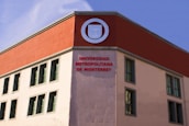 A building facade with a modern architectural design featuring a contrasting color scheme. The upper part of the structure is accented with a deep red section, prominently displaying a round emblem. Below, the words 'Universidad Metropolitana de Monterrey' are prominently written in red letters. The building has several rectangular windows with simple frames.