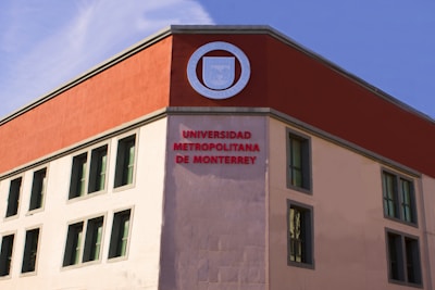 A building facade with a modern architectural design featuring a contrasting color scheme. The upper part of the structure is accented with a deep red section, prominently displaying a round emblem. Below, the words 'Universidad Metropolitana de Monterrey' are prominently written in red letters. The building has several rectangular windows with simple frames.