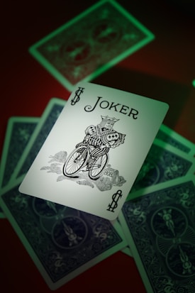 A Joker card prominently featuring a detailed illustration of a jester-like figure riding a bicycle, surrounded by partially visible playing cards with a back design. The lighting casts a greenish hue, adding an accent to the overall appearance.