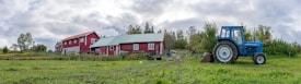 A rural scene featuring a farmhouse with red walls and a green roof, surrounded by open fields and trees under a cloudy sky. A blue tractor is parked on the grassy area near the house.