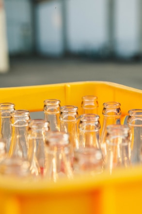 A group of empty clear glass bottles arranged within a bright yellow plastic crate, with a blurred industrial background.