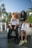 Two people are standing in front of a luxury car, both wearing sporty, casual outfits. One person has curly hair that is mid-motion, wearing a white t-shirt with 'SWAGG' and black pants. The other person smiles at the camera, wearing a black crop top with 'SWAGG' and white shorts. Lush palm trees and a clear blue sky form the backdrop, suggesting a sunny day.