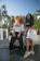 Two people are standing in front of a luxury car, both wearing sporty, casual outfits. One person has curly hair that is mid-motion, wearing a white t-shirt with 'SWAGG' and black pants. The other person smiles at the camera, wearing a black crop top with 'SWAGG' and white shorts. Lush palm trees and a clear blue sky form the backdrop, suggesting a sunny day.