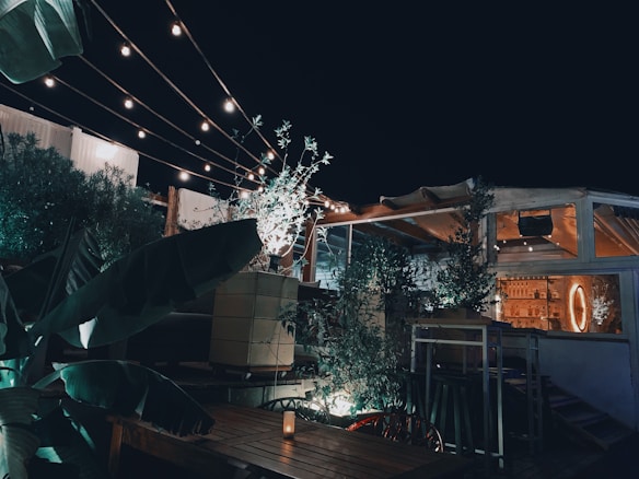 A cozy outdoor patio area is adorned with string lights that provide a warm and inviting ambiance. Wooden tables and chairs are neatly arranged under the lights, and lush green plants surround the space, creating a touch of nature. A dimly lit bar area is visible in the background, adding to the relaxed, intimate atmosphere of the setting.