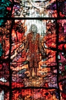A colorful stained glass artwork depicting a figure with flowing hair standing amidst a vibrant, abstract landscape. The glass is intricately designed with rich hues of red, orange, green, and purple, creating a sense of movement and complexity. The surrounding scenery includes abstracted forms resembling trees and a path leading into the distance.