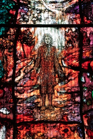 A colorful stained glass artwork depicting a figure with flowing hair standing amidst a vibrant, abstract landscape. The glass is intricately designed with rich hues of red, orange, green, and purple, creating a sense of movement and complexity. The surrounding scenery includes abstracted forms resembling trees and a path leading into the distance.