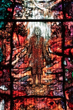 A colorful stained glass artwork depicting a figure with flowing hair standing amidst a vibrant, abstract landscape. The glass is intricately designed with rich hues of red, orange, green, and purple, creating a sense of movement and complexity. The surrounding scenery includes abstracted forms resembling trees and a path leading into the distance.