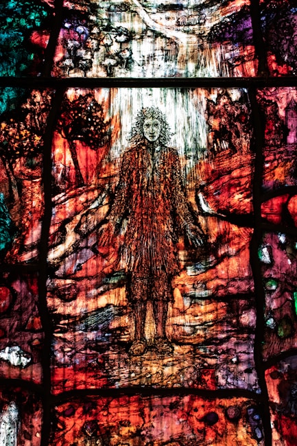 A colorful stained glass artwork depicting a figure with flowing hair standing amidst a vibrant, abstract landscape. The glass is intricately designed with rich hues of red, orange, green, and purple, creating a sense of movement and complexity. The surrounding scenery includes abstracted forms resembling trees and a path leading into the distance.