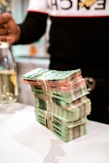 A large stack of banknotes is neatly arranged on a white surface, secured with rubber bands. In the background, a partially visible person is holding a bottle.