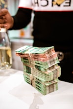 A large stack of banknotes is neatly arranged on a white surface, secured with rubber bands. In the background, a partially visible person is holding a bottle.