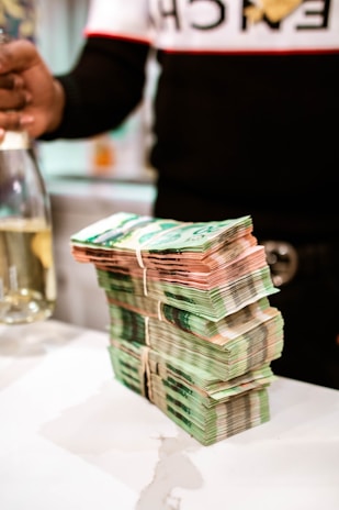 A large stack of banknotes is neatly arranged on a white surface, secured with rubber bands. In the background, a partially visible person is holding a bottle.