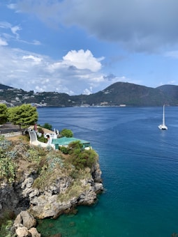 A serene coastal scene with a sailboat floating on clear blue waters. A rocky cliff adorned with lush greenery overlooks the water. A small white building with a green roof is situated near the edge of the cliff, with stairs leading down. In the background, hills with scattered buildings rise under a sky dotted with clouds.
