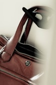 A close-up of a brown leather handbag with a zipper partially open and a circular logo badge. The handles are visible along with black sunglasses hanging on one of them.