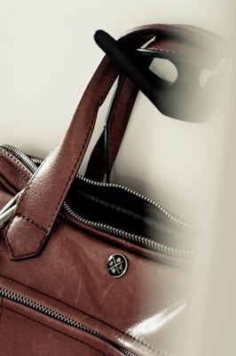 A close-up of a brown leather handbag with a zipper partially open and a circular logo badge. The handles are visible along with black sunglasses hanging on one of them.