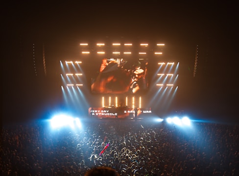 A large concert with an energetic crowd in front of a brightly lit stage. The stage features multiple LED screens displaying visuals and text, with powerful lights illuminating the audience. The atmosphere is lively and intense, reflecting a high-energy performance.
