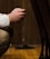 A person is kneeling or sitting with their hand holding a rosary bead string. The setting appears to be indoors, possibly in a church or a quiet, contemplative space. The focus is on the hand and the beads, with a wooden surface visible in the background.