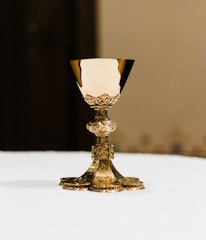 A detailed, ornate gold chalice is centered on a smooth white surface. The cup has elaborate engravings and a decorative pattern, with a wide base supporting the cup.
