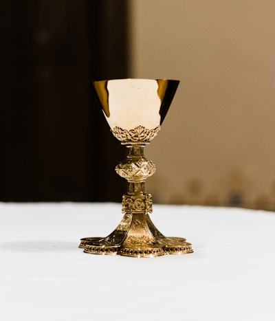 A detailed, ornate gold chalice is centered on a smooth white surface. The cup has elaborate engravings and a decorative pattern, with a wide base supporting the cup.